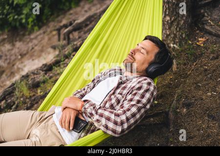 Man travels on bicycle, relaxing in green hammock, surfing Internet on smartphone, listening music on headphones in forest near Stock Photo