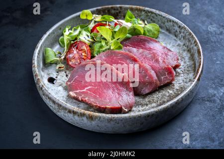 Modern style gourmet raw Japanese tataki beef filet with lettuce and tomatoes as close-up on a Nordic design plate with copy spa Stock Photo