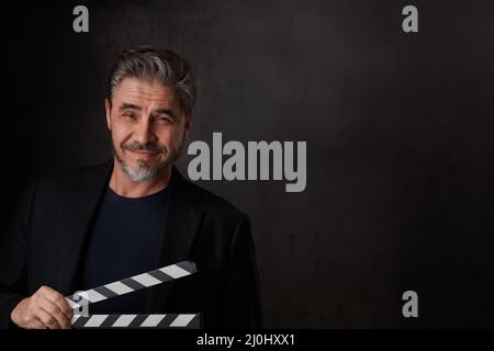 Happy man holding black clapper board. Movie director starting film. Copy space, dark background. Stock Photo