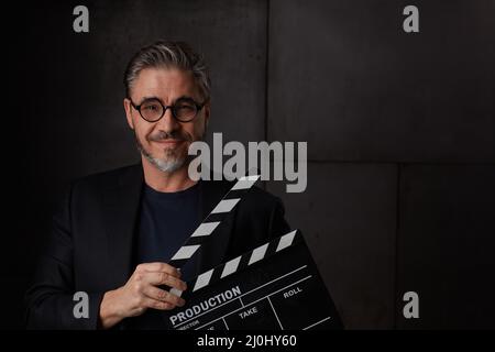 Happy man holding black clapper board. Movie director starting film. Copy space, dark background. Stock Photo