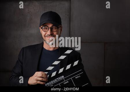Happy man holding black clapper board. Movie director starting film. Copy space, dark background. Stock Photo