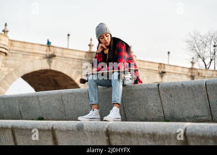 Young Hispanic woman sitting with skateboard on her knees. Venezuelan Latin woman in thought. Stock Photo