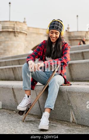 young woman sitting with skateboard. smiling latina listening to music. Stock Photo