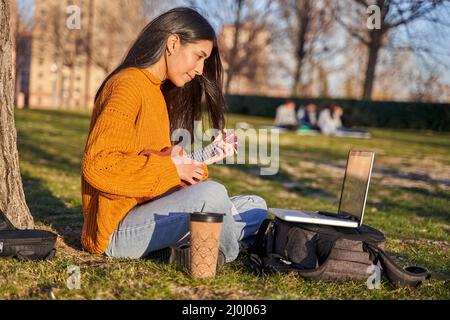 young latina playing the ukulele in a park. she takes a break from the city practicing in a park with her musical instrument, looking at the lyrics on Stock Photo