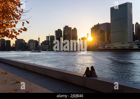 The sun sets among Midtown Manhattan skyscraper beyond the East River on November 2021 in New City. Two young women sit on the wall of Franklin D. Roo Stock Photo