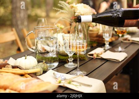 Glass of orange wine placed near dishware and bouquet of fresh flowers amidst assorted fruits on wooden table Stock Photo