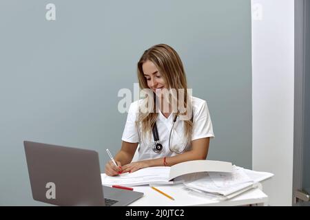 female doctor using laptop and writing notes in medical journal sitting at desk. Stock Photo