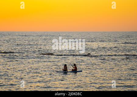 Two women sharing paddle board at sunset on calm waters in mediterranean sea in cyprus. Two girls on paddling on SUP. Silhuette Stock Photo