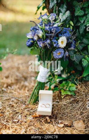 Wedding rings in a white box on dry grass with a bride's wedding bouquet of blue flowers. Stock Photo