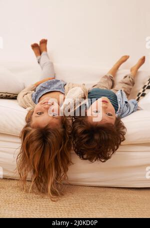 Even upside down were still bored. Portrait of two young children lying on a sofa with their heads hanging over the edge. Stock Photo