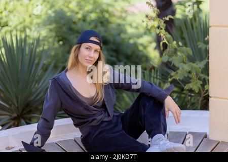Smiling blonde sports girl sitting on a wooden seat in the park against a background of green southern plants Stock Photo