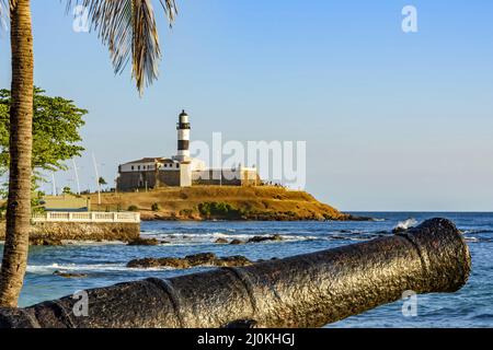 Old cannon and the Barra Lighthouse (Farol da Barra)  in the city of Salvador in Bahia surrounded by the sea Stock Photo