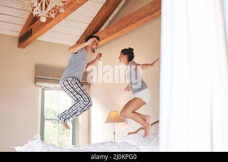 We bring a world of fun into each others lives. Full length shot of a cheerful young couple playing and jumping on their bed in their bedroom at home. Stock Photo