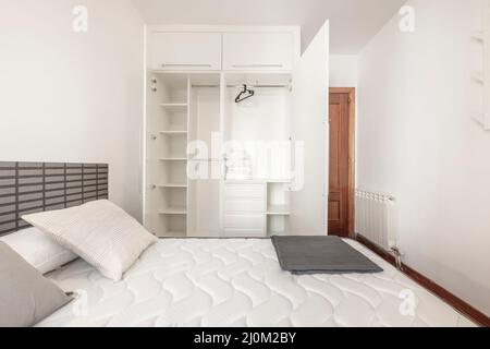Open white wardrobe with shelves, drawers, chest, clothes rail and black hangers in bedroom with king-size mattress and aluminum radiator Stock Photo