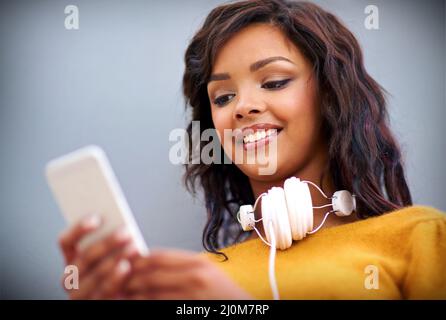 Her friend just sent her a song to listen to. Studio shot of a young woman with headphones around her neck using a mobile phone against a gray Stock Photo