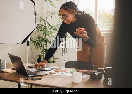 Freelancer taking a phone call in her home office. Happy female photographer smiling cheerfully while using a laptop. Creative young woman communicati Stock Photo