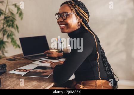 Happy female photographer smiling cheerfully in her home office. Creative young woman sitting at a desk with photographic equipment. Creative female f Stock Photo