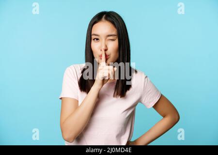 Smiling coquettish asian girl shushing, making silence taboo gesture, winking and making hush sign, press finger to lips, standi Stock Photo