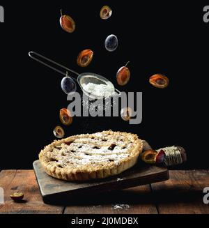 Baked round pie with plums on a levitating wooden kitchen board, top of a sieve with powdered sugar and livitating slices of plu Stock Photo