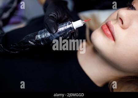 Cosmetologist applying red permanent make up tattoo on young woman lips. Permanent lips tattoo procedure concept. Stock Photo