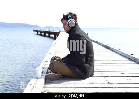 Young gloomy man listening to music sitting all alone on pier on a windy spring day. Stock Photo