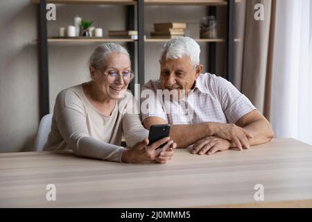 Smiling older couple sit at table using modern smartphone Stock Photo