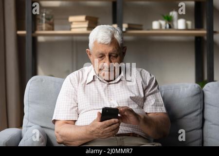 Serious older man sit on sofa with modern smartphone Stock Photo