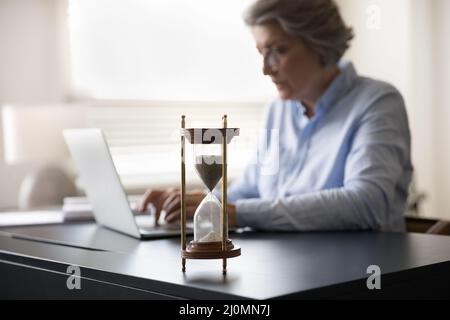 Mature businesswoman work on laptop, hourglass on table, forefront view Stock Photo