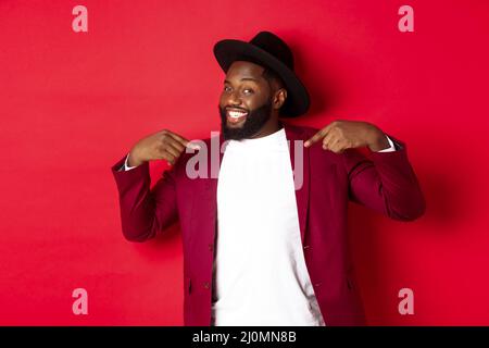 Christmas shopping and people concept. Handsome Black man pointing at himself, going on party, standing confident against red ba Stock Photo