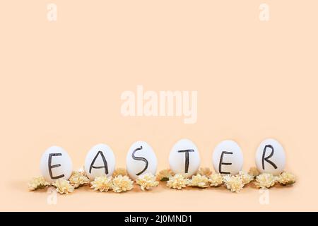 White eggs with black letters EASTER around flowers on yellow background. Happy Easter card. Flat lay, top view, copy space. Stock Photo