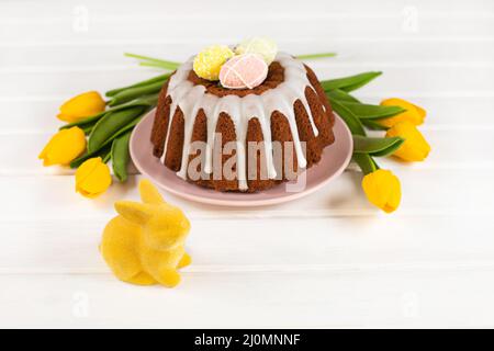 Glazed easter cake decorated with mini eggs candy, yellow tulips and Easter Bunny on white table. Happy Easter Holidays. Stock Photo