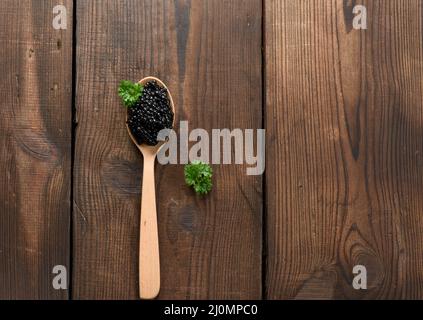 Fresh grainy black paddlefish caviar in brown wooden spoon on a black background, top view Stock Photo