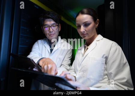 Diverse male and female engineers wearing aprons discussing over clipboard in computer server room Stock Photo