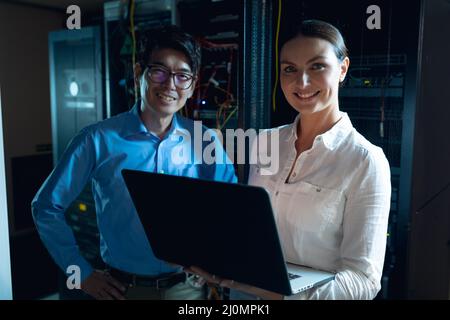 Portrait of diverse male and female engineers with laptop smiling in computer server room Stock Photo