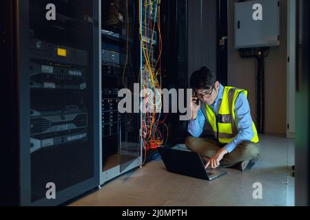 Asian male engineer using laptop and talking on smartphone while inspecting in computer server room Stock Photo