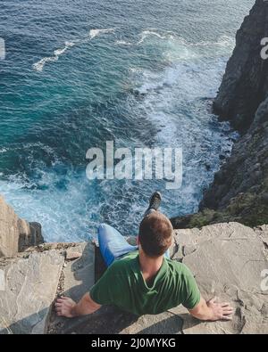 Top view of a man in green shirt sitting on the edge of the cliff with dramatic sea view Stock Photo