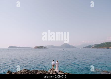 Bride and groom stand on the rocky shore holding hands looking at sea Stock Photo