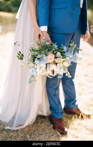 Groom in a blue suit holds a bouquet of flowers in his hand. Bride in a white dress hugs him from behind. Close-up Stock Photo