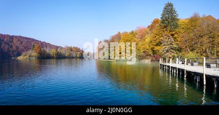 Horizontal view of calm lake with a waterfall and colorful fall foliage and a dock in the foreground Stock Photo