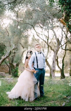 The bride and groom are standing among the trees in the olive grove, the bride hugging the groom from behind Stock Photo