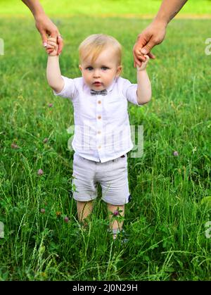 Baby child on the green grass in summer park. Parents hand and child. Perfect family holding hands, adopted child being supported by loving parents. Stock Photo