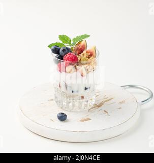 Transparent glass with granola poured with yogurt, on top of ripe raspberries, blueberries and figs on a white table. Healthy br Stock Photo