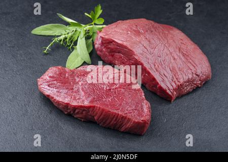 Raw dry aged bison beef rump steak piece and slices with herbs offered as close-up on black background Stock Photo