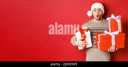 Concept of winter holidays, new year and celebration. Image of surprised handsome guy in sweater and santa hat, receiving gifts, Stock Photo