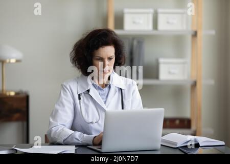 Serious senior chief doctor woman using laptop at workplace Stock Photo