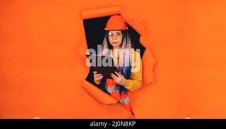 Female construction worker holding clipboard and sticking out of hole of orange background. Young woman in eyeglasses and overalls checking business p Stock Photo