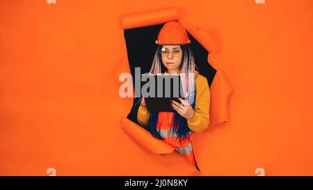Female construction worker holding clipboard and sticking out of hole of orange background. Young woman in eyeglasses and overalls checking business p Stock Photo
