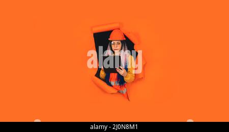 Female construction worker holding clipboard and sticking out of hole of orange background. Young woman in eyeglasses and overalls checking business p Stock Photo