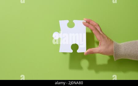 Female hand holds a fragment of a white large puzzle on a green background, the concept of finding an idea, solving a problem Stock Photo