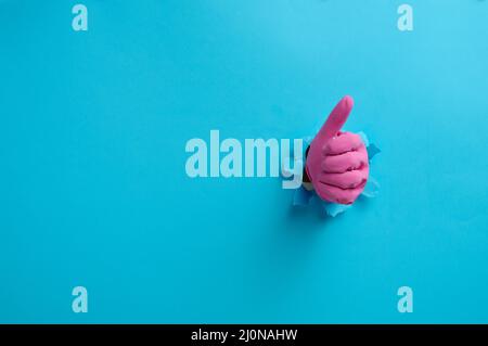 A hand in a pink household rubber glove sticks out of the torn hole and shows a gesture okay, thumb up. Blue background Stock Photo